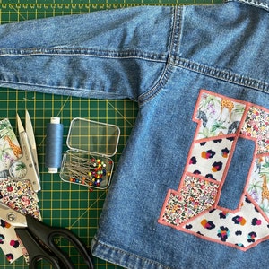 May include: A blue denim jacket with a large letter 'D' appliquéd on the back, made from patterned fabric patches. Sewing tools, including scissors, pins, and thread, are arranged around the jacket on a green cutting mat.