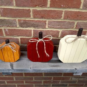 May include: Three wooden pumpkin decorations in orange, red, and cream. Each pumpkin features vertical paneling, a dark brown stem, and a tied jute twine bow. The pumpkins are arranged in a row, varying in size from smallest to largest.