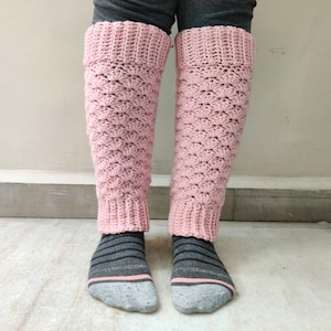 May include: A pair of pink crocheted leg warmers with a textured pattern. The leg warmers are worn on a person's legs.