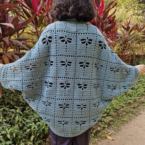 Könnte beinhalten: Hellblauer gehäkelter Schal mit Libellenmuster. Der Schal ist aus weichem Garn gefertigt und hat ein lockeres, fließendes Design. Die Libellen-Ausschnitte verleihen dem Kleidungsstück eine dekorative Note.