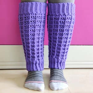 May include: Purple crocheted leg warmers with a textured pattern. The leg warmers are worn over gray and pink socks.