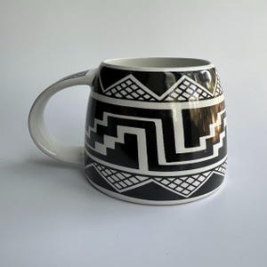 May include: A ceramic mug featuring a high-contrast black and white geometric pattern. The design includes a central stepped motif flanked by triangular sections with a cross-hatched grid texture. The handle and interior are solid white.