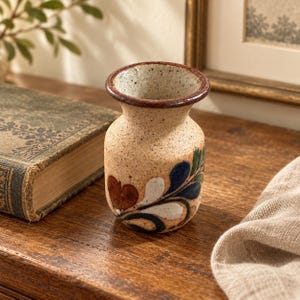 May include: A small, speckled beige ceramic vase with a flared rim and a dark brown glazed edge. The body features hand-painted floral motifs in shades of deep blue, green, white, and reddish-brown, arranged in a curved, leafy pattern.
