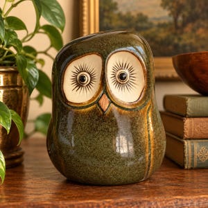 May include: A ceramic owl figurine with a glossy, mottled dark green glaze. The owl features two large, cream-colored circular eye patches with radiating dark lines, black pupils, and a small, pointed, light brown beak centered on the face.