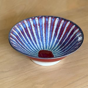 Può includere: Una ciotola in ceramica con un design radiale nei toni del blu, rosso e bianco. La ciotola ha una base rossa e un bordo bianco. L'interno presenta un centro rosso scuro. Questa ciotola decorativa è adatta per servire cibo.
