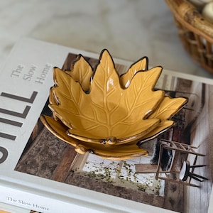 May include: Two nested ceramic bowls shaped like maple leaves in a mustard yellow glaze with dark brown edges. The bowls feature embossed vein details and are stacked on top of a book titled The Slow Home by Natalie Walton with a white cover.