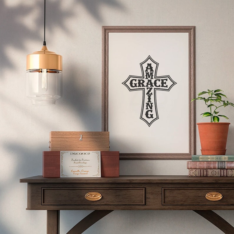 May include: A framed print with a black cross design that reads "AMAZING GRACE" on a white background. The print is hanging on a wall above a wooden console table with two drawers. A wooden box with a label that reads "Duccance" is on the table. A gold pendant light hangs above the table.