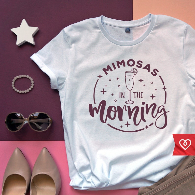 May include: A light blue t-shirt with a maroon circle graphic that says "Mimosas in the morning" with a stylized illustration of a glass of champagne with a lemon slice.