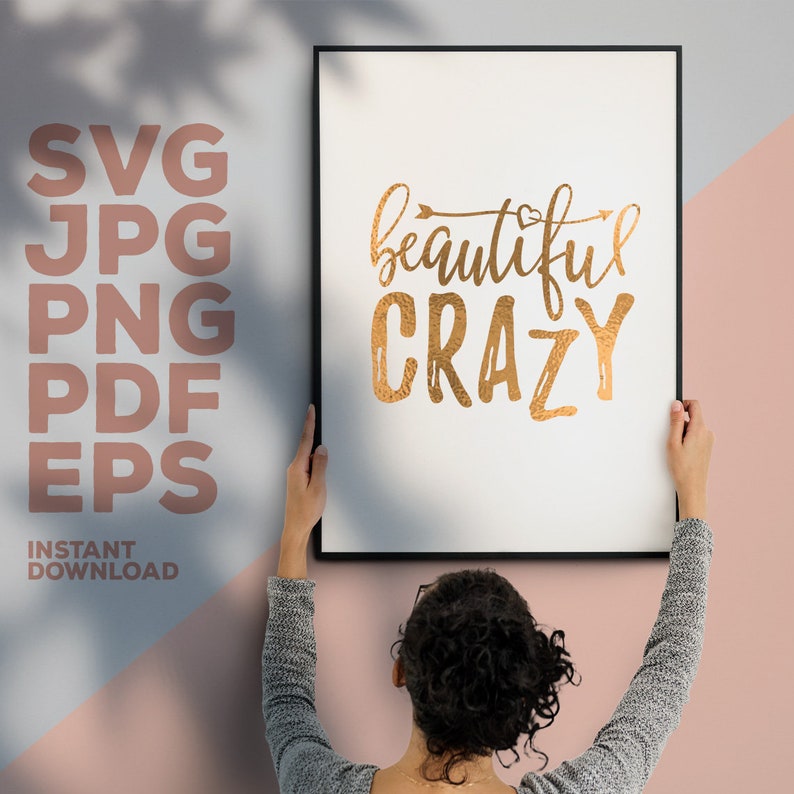 May include: A gold foil print with the text "beautiful crazy" on a white background in a black frame. The print is being hung on a pink wall.
