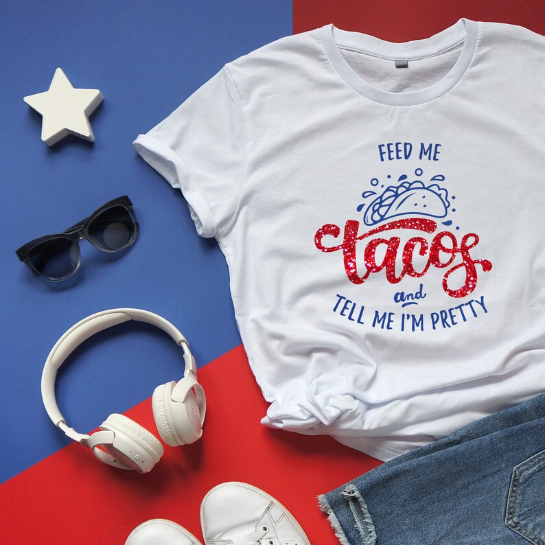 May include: White t-shirt with the text "Feed me tacos and tell me I'm pretty" in red and blue. The shirt is styled with sunglasses, white headphones, a star, white sneakers, and denim shorts on a red and blue background.
