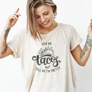 May include: A beige t-shirt with the text "Feed me tacos and tell me I'm pretty" in dark green. The graphic includes a taco illustration. The shirt is made of a soft material.