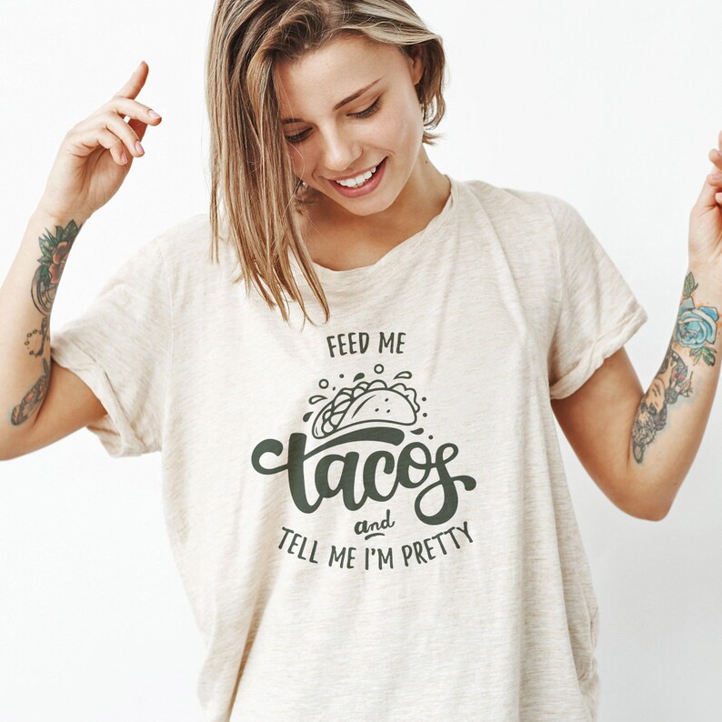 May include: A beige t-shirt with the text "Feed me tacos and tell me I'm pretty" in dark green. The graphic includes a taco illustration. The shirt is made of a soft material.