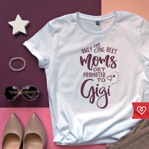 Może przedstawiać: Jasno błękitny t-shirt z napisem "Only the best moms get promoted to Gigi" w kolorze bordowym z dwoma małymi sercami.