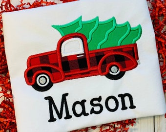 May include: A white t-shirt featuring an embroidered red and black buffalo plaid truck carrying a green Christmas tree. The name Mason is embroidered in black lettering below the truck design. The shirt is displayed on a white wooden surface.