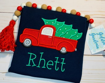 May include: A navy blue t-shirt featuring an embroidered red pickup truck carrying a green Christmas tree. The name Rhett is embroidered in teal below the truck. A string of red, white, and natural wood beads with a red tassel is attached.