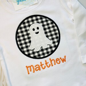 May include: White long-sleeved shirt with a black and white checkered circle containing a white ghost with a smiling face. The name "matthew" is embroidered in orange below the circle.