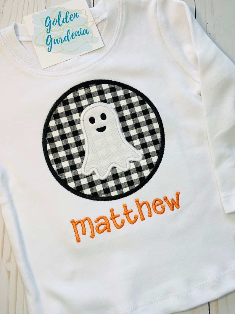 May include: White long-sleeved shirt with a black and white checkered circle containing a white ghost with a smiling face. The name "matthew" is embroidered in orange below the circle.