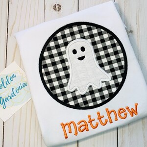 May include: White t-shirt with a black and white checkered circle with a white ghost inside. The name "matthew" is embroidered in orange below the circle.