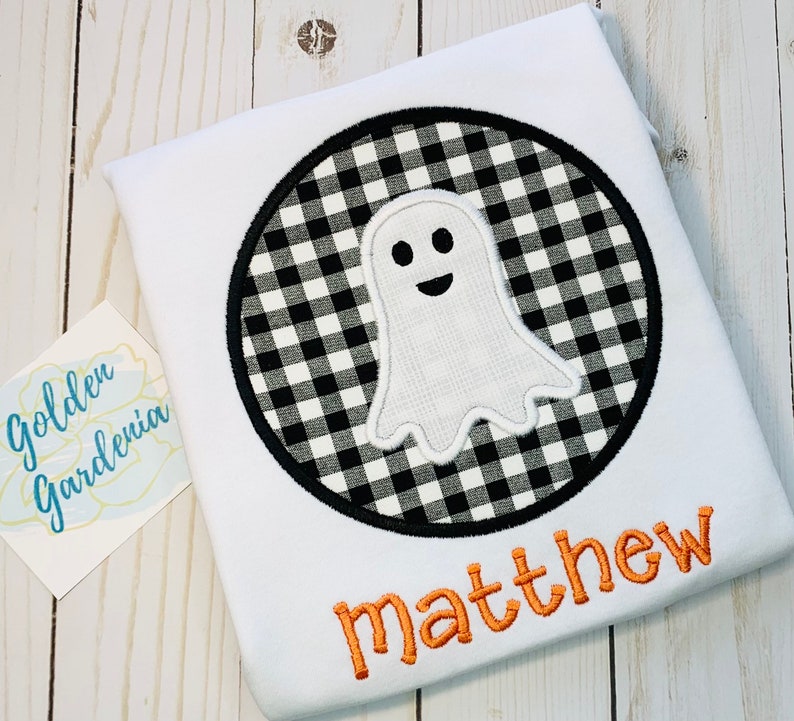 May include: White t-shirt with a black and white checkered circle with a white ghost inside. The name "matthew" is embroidered in orange below the circle.