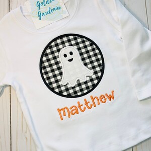 May include: White long-sleeved shirt with a black and white checkered ghost applique in a circle. The name "matthew" is embroidered in orange below the ghost.