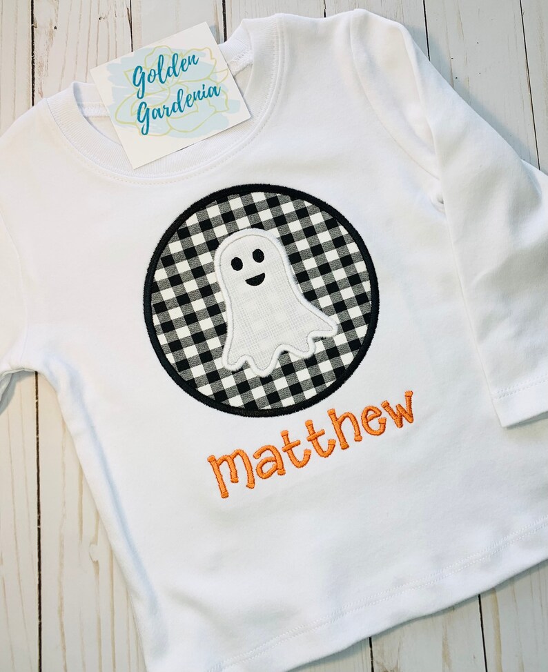 May include: White long-sleeved shirt with a black and white checkered ghost applique in a circle. The name "matthew" is embroidered in orange below the ghost.