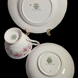 May include: A white teacup, saucer, and plate set with a floral design. The teacup and saucer have pink flowers. The plate and saucer have a scalloped edge. The set is marked "Tuscan Fine English Bone China".
