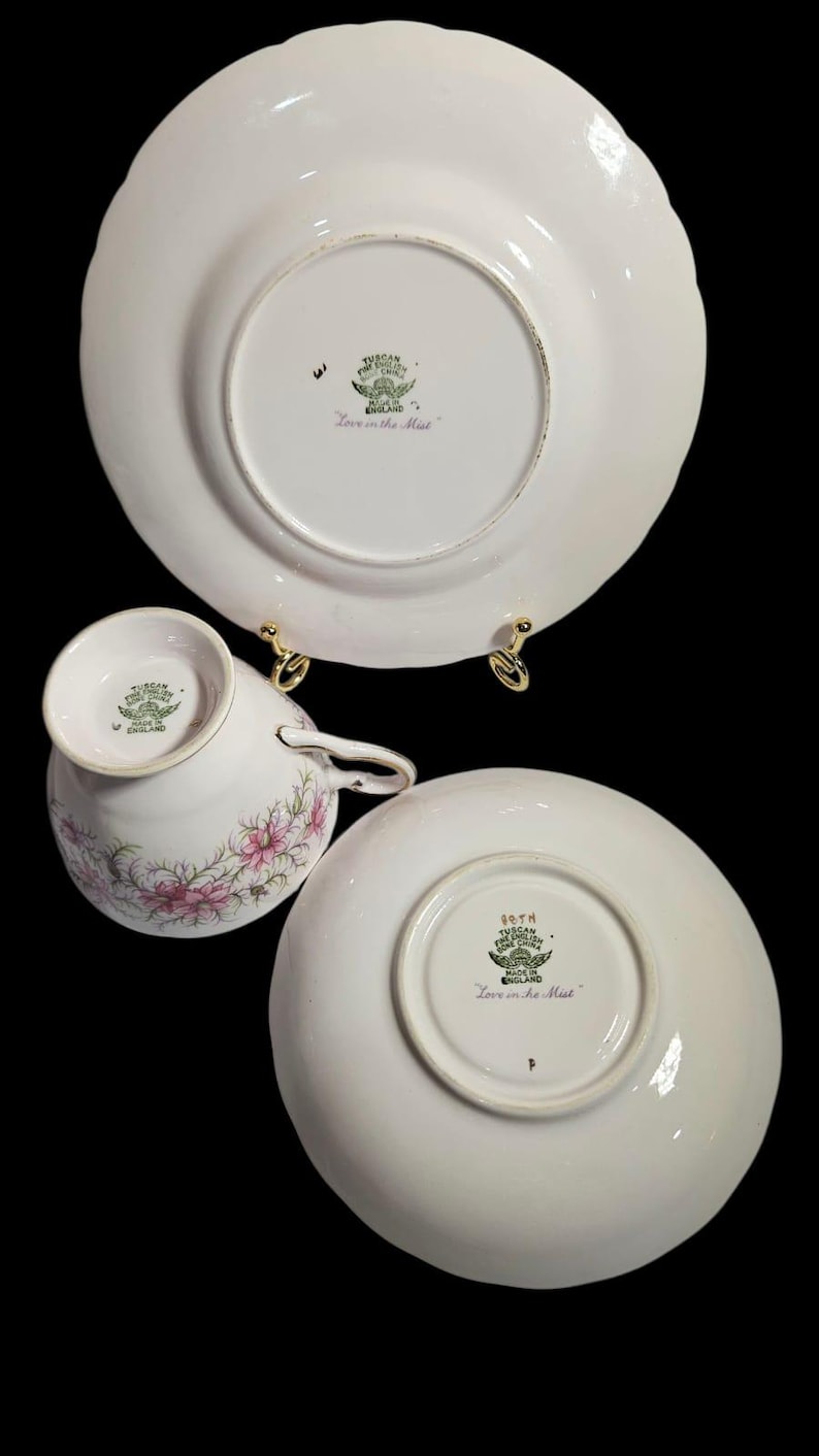 May include: A white teacup, saucer, and plate set with a floral design. The teacup and saucer have pink flowers. The plate and saucer have a scalloped edge. The set is marked "Tuscan Fine English Bone China".