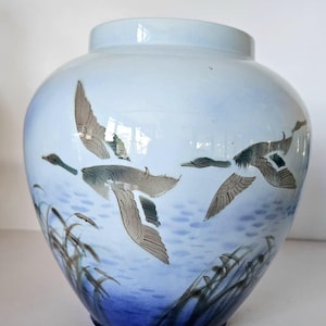May include: A light blue ceramic vase with a design of flying ducks over water. The ducks are depicted in shades of gray and brown, with detailed wings. The water and sky are painted in shades of blue, with reeds at the base.