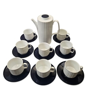 May include: A white ceramic coffee set with a ribbed texture, including a tall coffee pot, eight cups, and saucers. The saucers are a dark navy blue. The set is designed for serving coffee or tea.