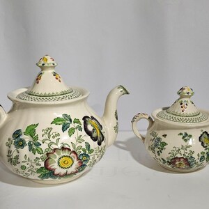 May include: A white teapot and sugar bowl with floral designs in shades of green, yellow, and pink. The teapot has a lid with a floral design and a spout. The sugar bowl has a lid with a floral design and a handle.