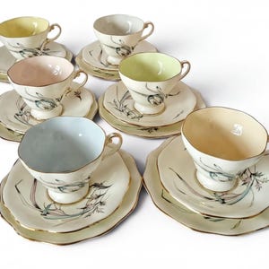 May include: A set of six porcelain teacups and saucers featuring pastel-colored interiors in yellow, pink, light green, and blue. Each piece is decorated with a delicate botanical illustration of green stems and leaves, finished with gold trim.
