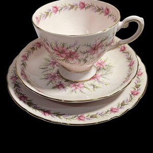 May include: A vintage teacup, saucer, and side plate set. The set features a pink floral pattern with gold trim on a white background. The teacup sits atop the saucer, with a side plate below. The set is likely porcelain.