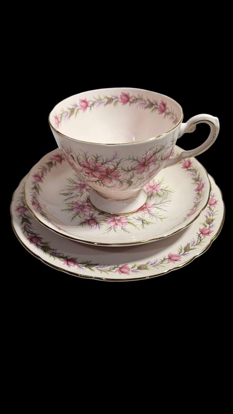 May include: A vintage teacup, saucer, and side plate set. The set features a pink floral pattern with gold trim on a white background. The teacup sits atop the saucer, with a side plate below. The set is likely porcelain.