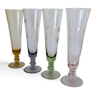 May include: A set of four tall, slender glass flutes, each featuring a unique pastel-coloured base in amber, purple, green, and pink. The clear upper bowls are decorated with delicate, etched starburst patterns, and each glass stands on a round base.