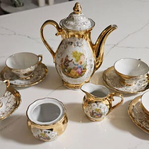 Puede incluir: Un juego de té de porcelana vintage con ribetes dorados. El juego incluye una tetera, una jarra de crema, un azucarero y varias tazas de té con platillos. Cada pieza presenta un fondo blanco con una escena decorativa y detalles dorados.