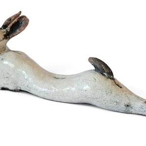 May include: A ceramic hare figurine in a prone, stretched-out position. The sculpture features a glossy, off-white glazed finish with dark, weathered-looking accents on the ears, nose, tail, and hind legs, creating a textured, rustic appearance.