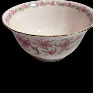 May include: A white porcelain bowl with a gold rim and a floral pattern. The bowl features pink flowers and green leaves. The rim of the bowl is also decorated with a floral pattern. The bowl is set against a black background.