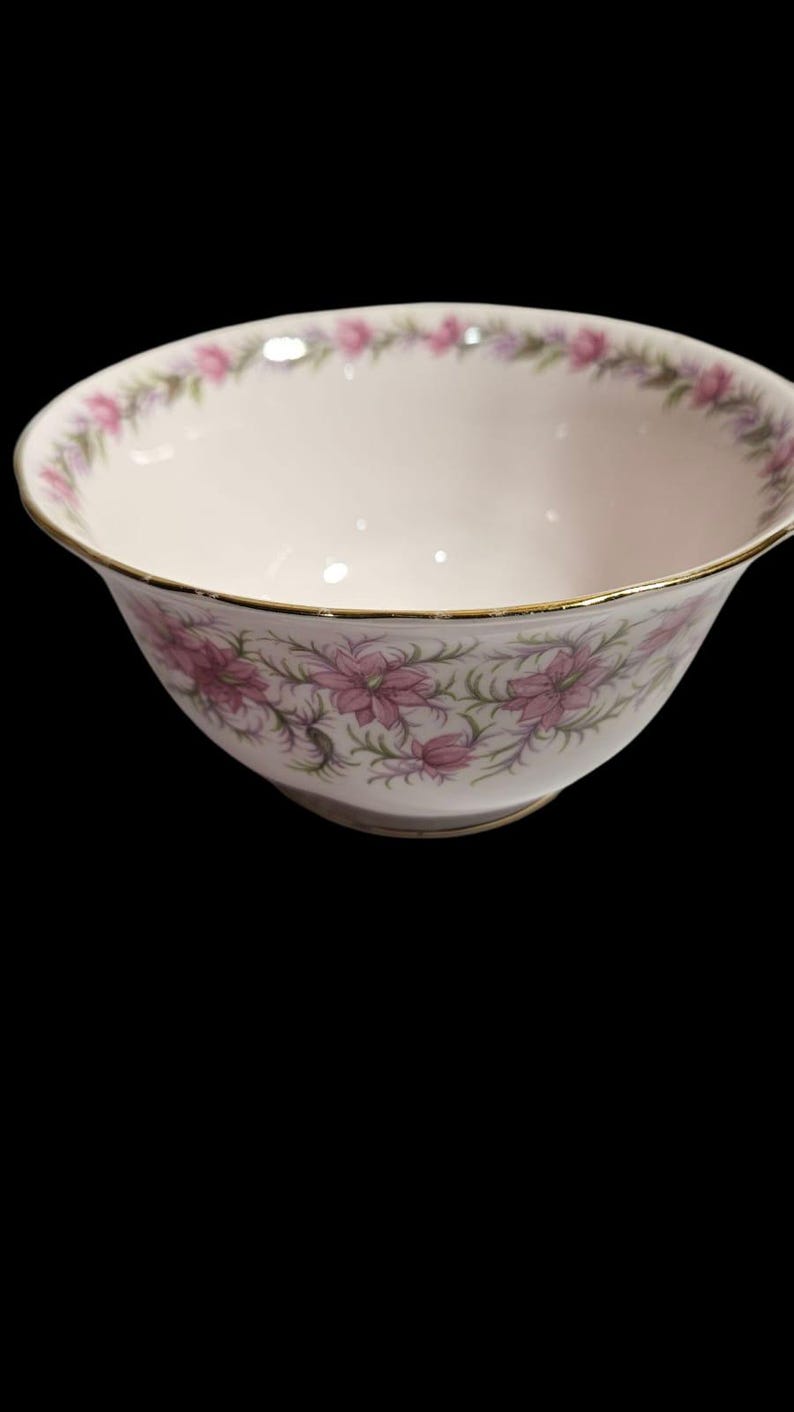 May include: A white porcelain bowl with a gold rim and a floral pattern. The bowl features pink flowers and green leaves. The rim of the bowl is also decorated with a floral pattern. The bowl is set against a black background.