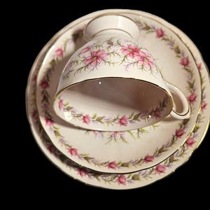 May include: A vintage teacup, saucer, and dessert plate set. The white porcelain pieces feature a pink floral design with gold trim. The teacup is resting on the saucer, and the dessert plate is below.