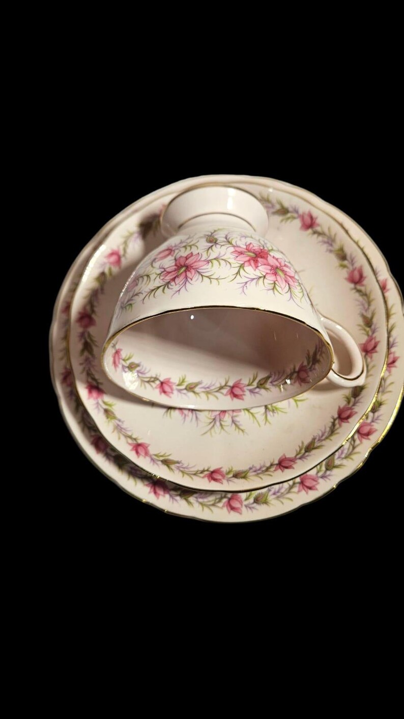 May include: A vintage teacup, saucer, and dessert plate set. The white porcelain pieces feature a pink floral design with gold trim. The teacup is resting on the saucer, and the dessert plate is below.
