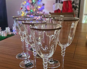 May include: A set of six clear wine glasses with a decorative etched design and a gold rim. The glasses are displayed on a wooden table. The background includes a Christmas tree with colorful lights and holiday decorations.