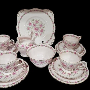 May include: A vintage tea set with a floral design. The set includes teacups, saucers, a serving bowl, a creamer, and a serving plate. The pieces are white with pink floral patterns and gold trim. The set is arranged on a black background.