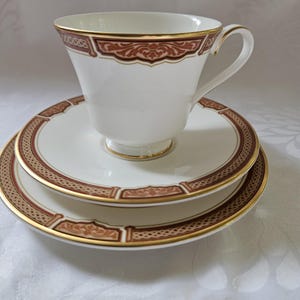Puede incluir: Un juego de taza y platillo de té blanco con borde dorado y un patrón floral marrón y dorado.