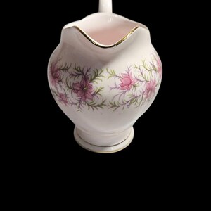 May include: A white ceramic creamer with a pink interior and a gold rim. The creamer features a floral design with pink flowers and green leaves. The creamer has a small handle and a decorative base. The background is black.