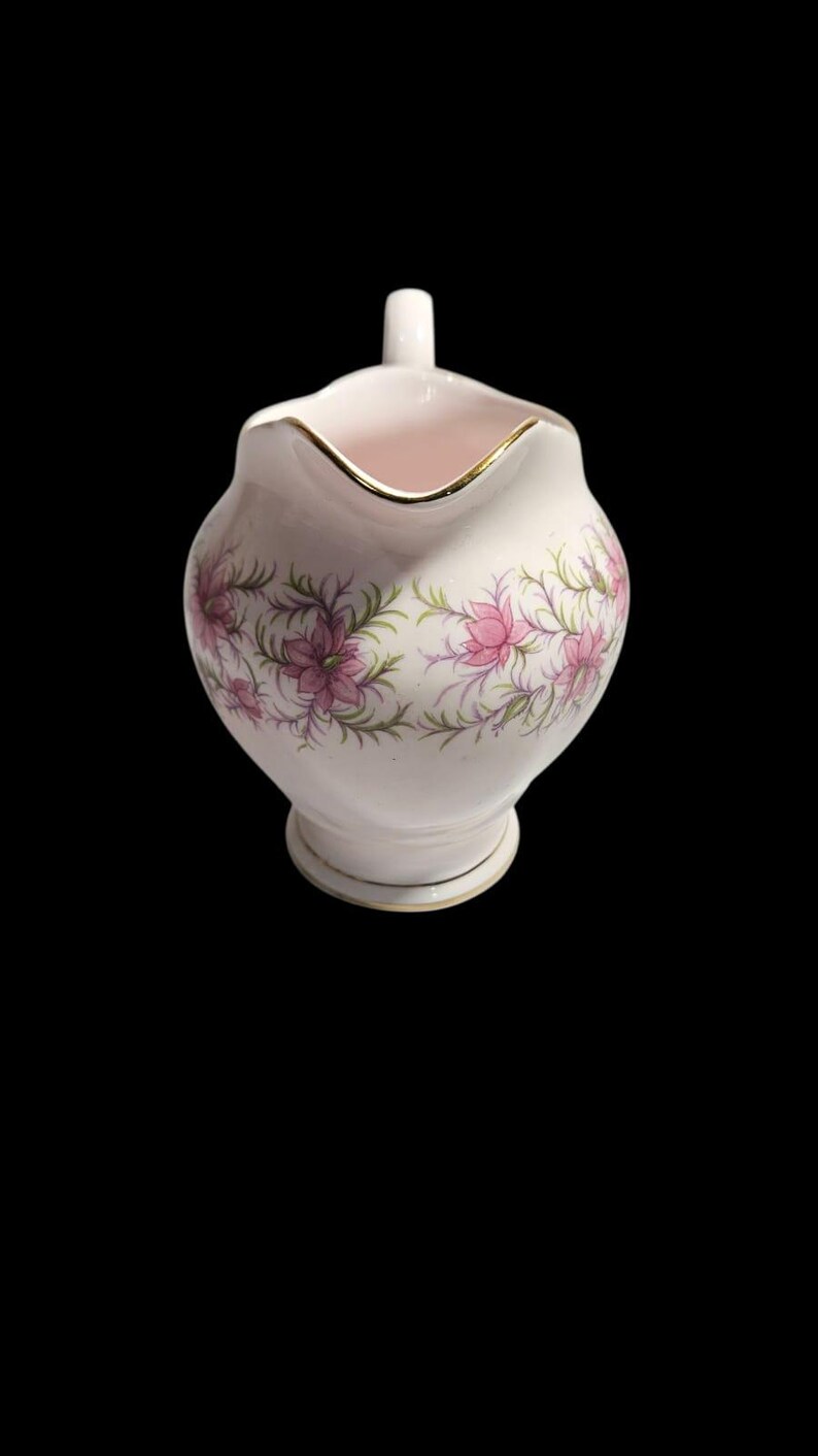 May include: A white ceramic creamer with a pink interior and a gold rim. The creamer features a floral design with pink flowers and green leaves. The creamer has a small handle and a decorative base. The background is black.