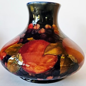 Puede incluir: Un jarrón decorativo de cerámica con acabado brillante. El jarrón presenta un borde azul oscuro y un cuerpo adornado con un diseño colorido de frutas y hojas en tonos rojos, naranjas, dorados y morados. El jarrón tiene forma bulbosa.
