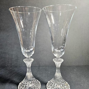 May include: A pair of clear glass champagne flutes featuring smooth, flared bowls and intricate, vertically ribbed stems that transition into wide, textured circular bases with a radial pattern, providing a stable foundation for each glass.