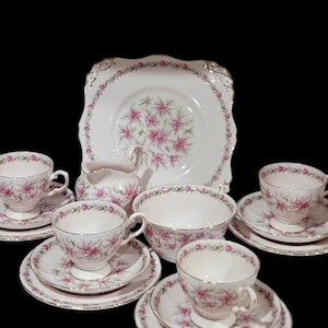 May include: A vintage tea set with a floral pattern. The set includes teacups, saucers, a sugar bowl, a serving bowl, and a cake plate. The pieces are white with pink floral designs and gold trim. A classic tea party set.