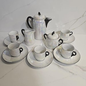 Puede incluir: Un juego de té iridiscente vintage con un acabado nacarado. El juego incluye una tetera, una jarra de crema, un azucarero y seis tazas de té con platillos. Cada pieza tiene detalles negros en las asas y los bordes.