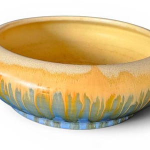 May include: A round ceramic bowl featuring a smooth, wide rim with a yellow glaze that transitions into a textured, dripping orange and blue glaze pattern on the exterior. The interior of the bowl is finished in a solid, pale yellow glaze.